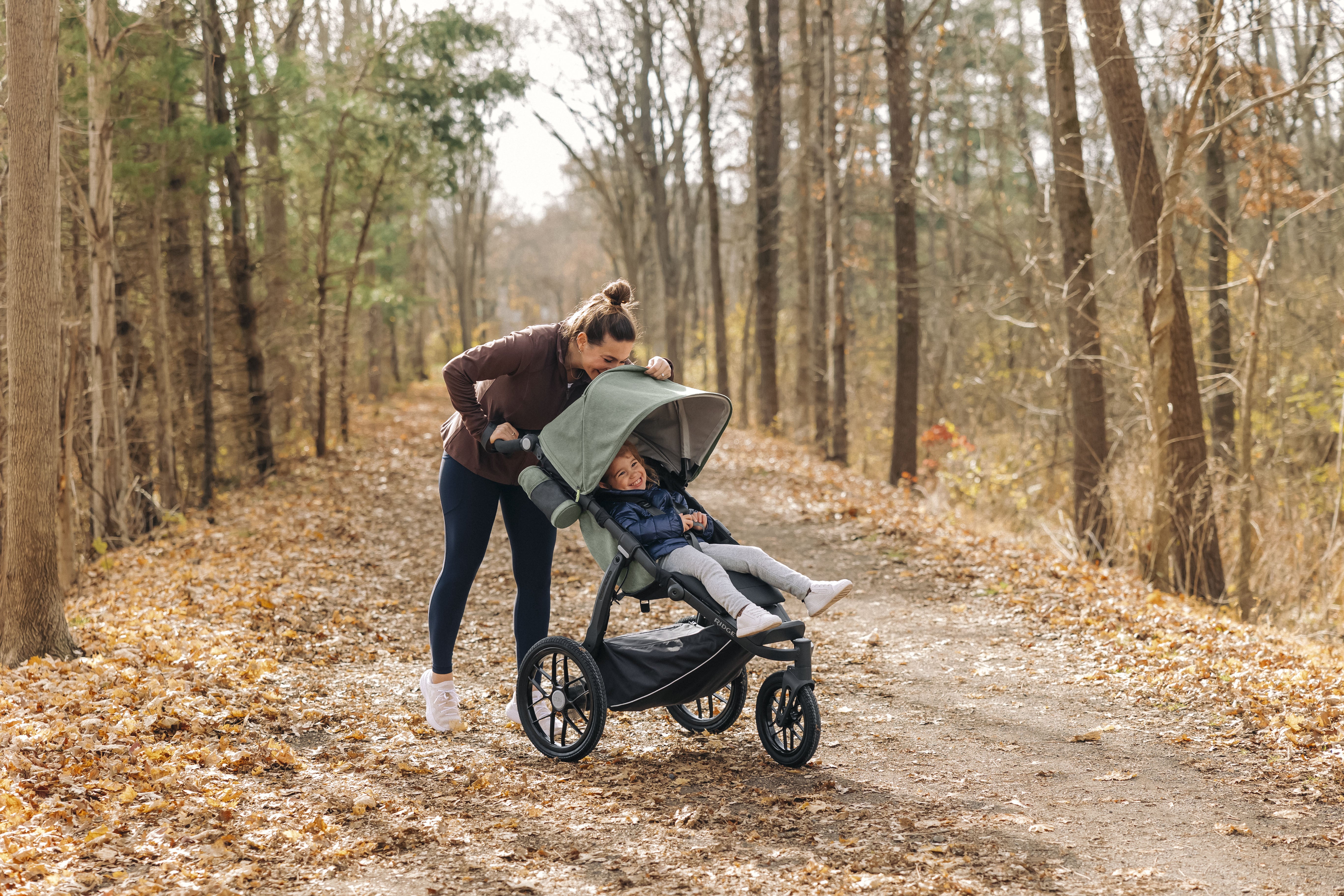A woman strolls through the woods, pushing a baby in the UPPAbaby Ridge stroller, surrounded by lush greenery and tall trees.