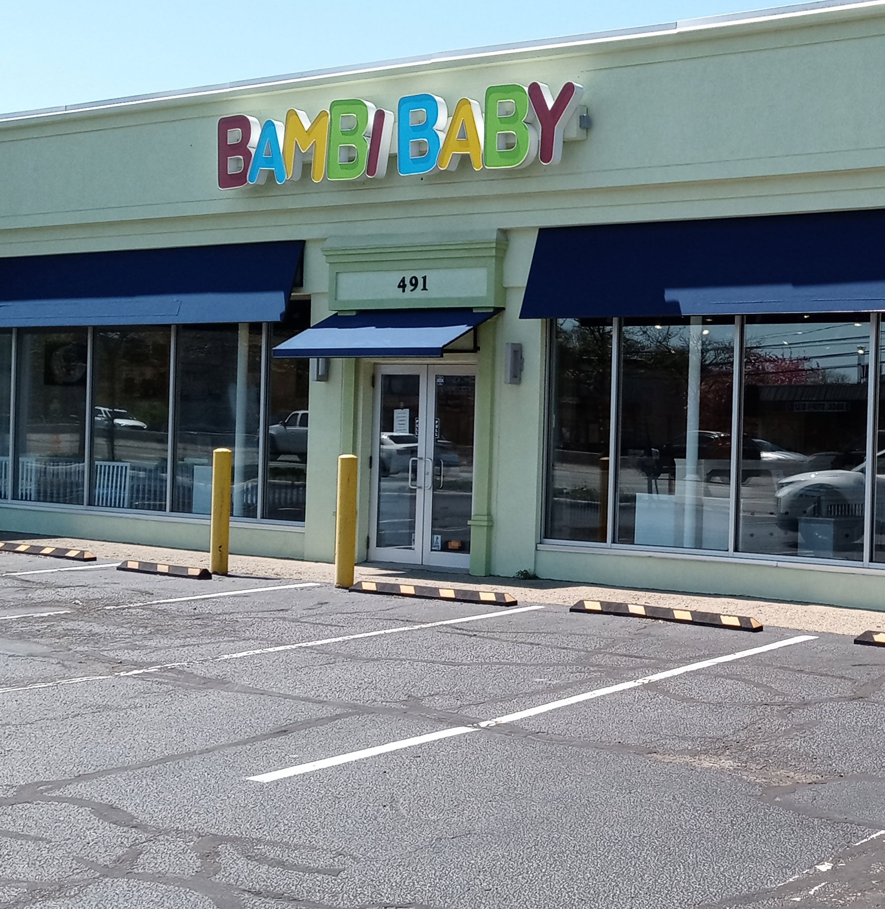 A storefront with blue awnings, displaying the sign "Bambi Baby" and the number 491 above the entrance. The parking lot in front is empty.