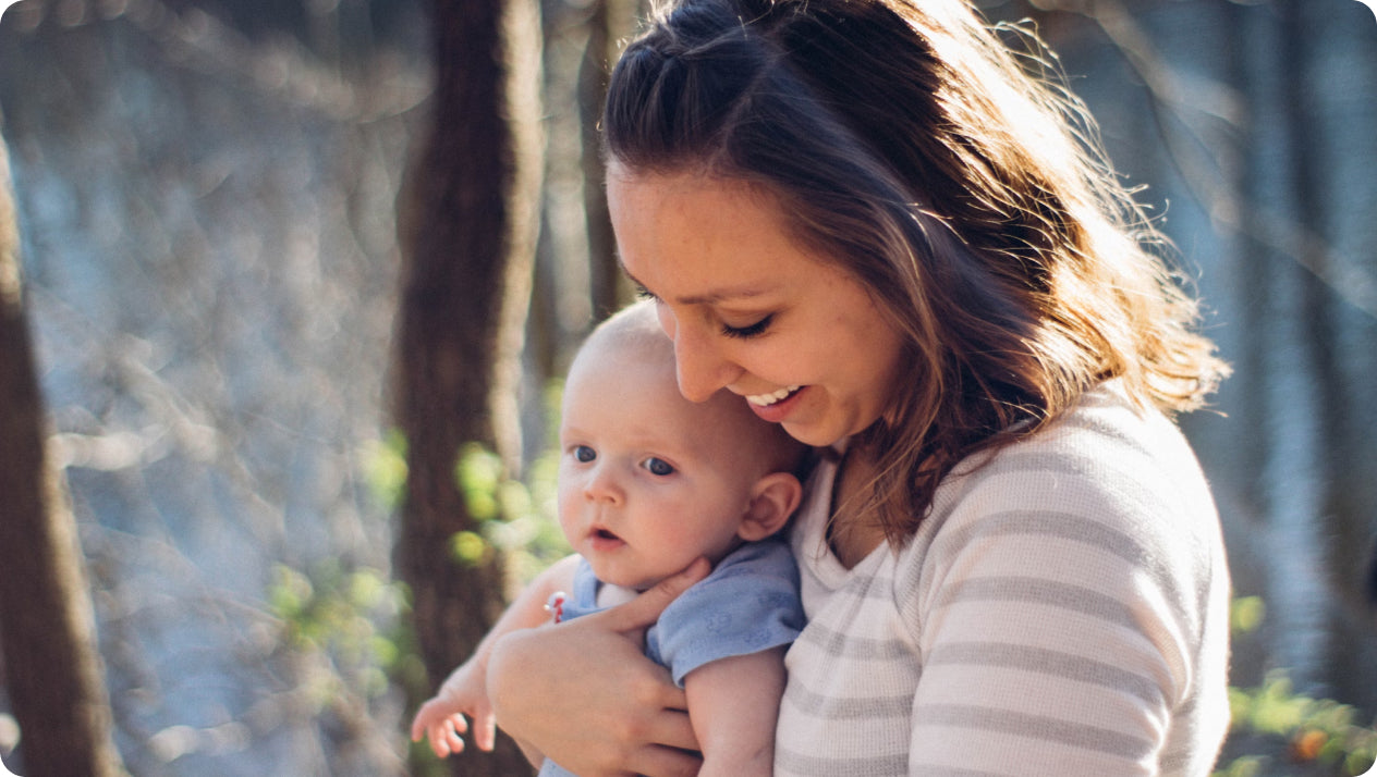 A woman with shoulder-length brown hair, wearing a light-striped shirt, holds a baby in her arms. Both are outdoors, with trees and sunlight in the background. The woman is smiling and looking down at the baby, who has a curious expression.