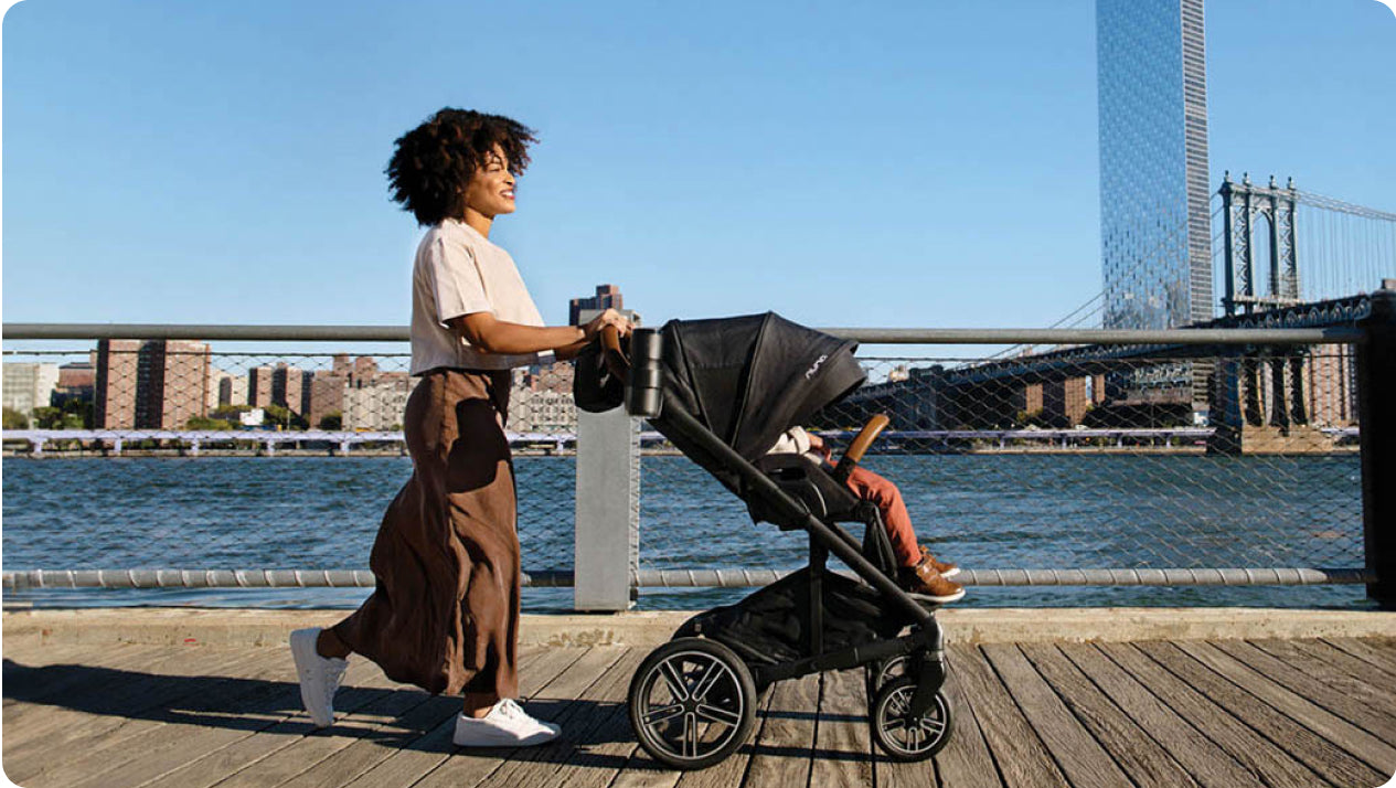 A person walks along a wooden promenade by a river, pushing a stroller with a child. The background features a cityscape with high-rise buildings and a bridge under a clear blue sky. The person wears a light-colored top and brown pants while the child is seated in the stroller.