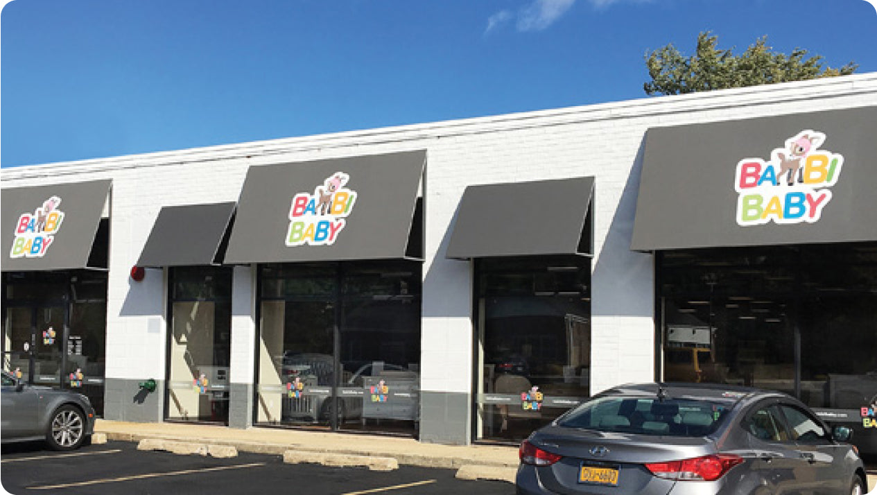 A storefront with three large windows and gray awnings displaying the "Bambi Baby" logo. Several parked cars are in front of the store, and the sky is clear with a few scattered clouds.