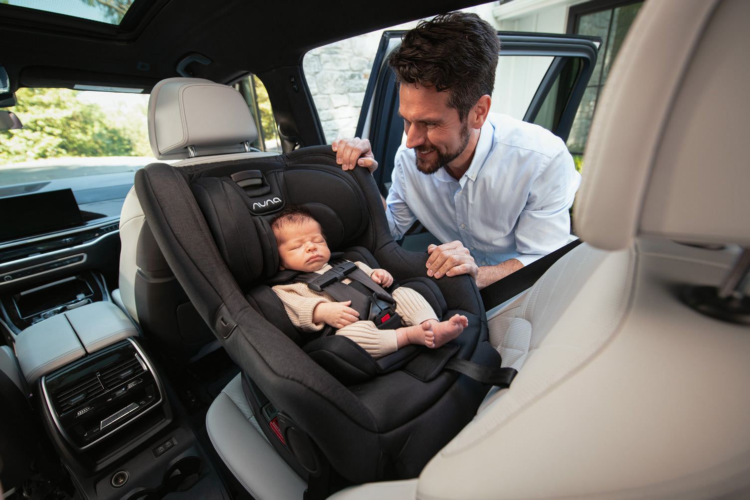 A man is adjusting a sleeping baby in the Nuna Rava car seat inside a vehicle.