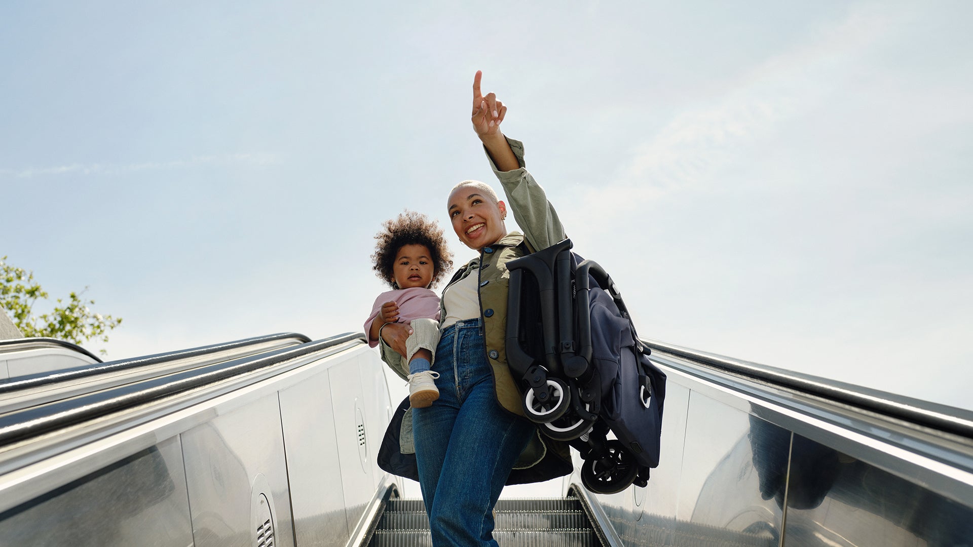 A woman and a child are riding an escalator together, enjoying their time as they move between floors, with the woman carrying the folded Bugaboo Butterfly stroller.