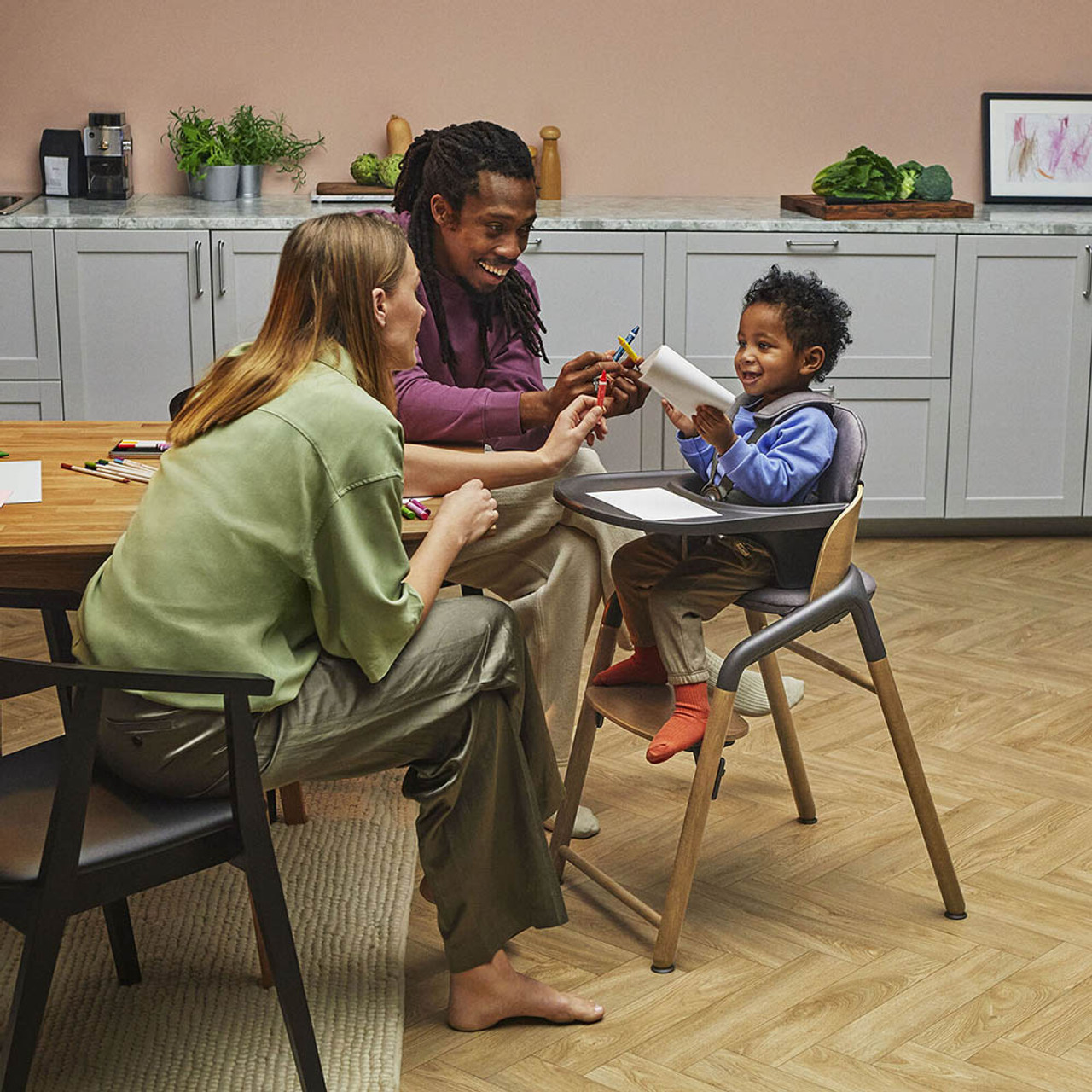 Three people sitting at a kitchen table, with an adult helping a toddler use a pair of scissors.