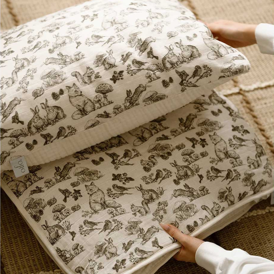 Two hands holding a stack of pillows with intricate nature-themed illustrations on the fabric, featuring animals and plants. The background shows a textured rug.