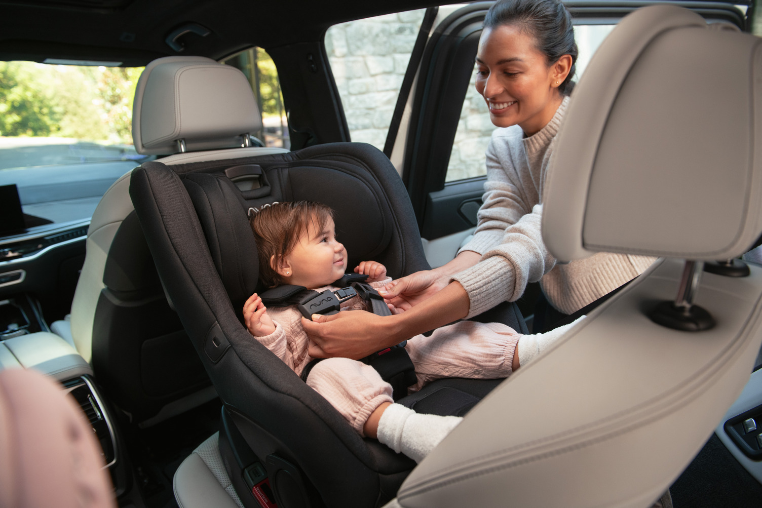 A toddler sits safely in a rear-facing car seat inside a vehicle, smiling and looking to the side.